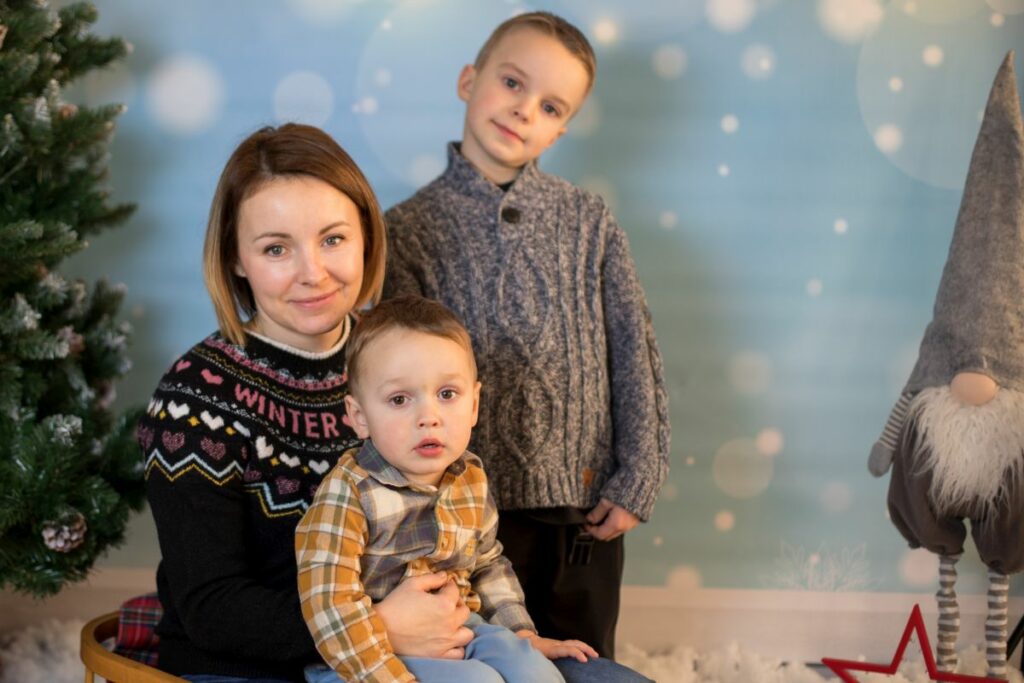 Na zdjęciu widoczna jest mama oraz dwóch synów podczas mini sesji świątecznej. Mama z młodszym chłopcem siedzi na sankach, a starszy z chłopców stoi obok nich. W tle choinka, szary skrzat oraz zimowe tło