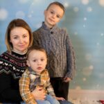 Na zdjęciu widoczna jest mama oraz dwóch synów podczas mini sesji świątecznej. Mama z młodszym chłopcem siedzi na sankach, a starszy z chłopców stoi obok nich. W tle choinka, szary skrzat oraz zimowe tło