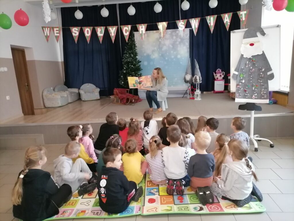 W sali na podium siedzi pani bibliotekarka i czyta książkę dzieciom siedzącym przed podium. Z boku po prawej stronie duży mikołaj biało szary. Z tyłu za pania bibliotekarką granatowe zasłony, choinka, sanki oraz trzy fotele.