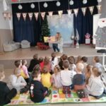 W sali na podium siedzi pani bibliotekarka i czyta książkę dzieciom siedzącym przed podium. Z boku po prawej stronie duży mikołaj biało szary. Z tyłu za pania bibliotekarką granatowe zasłony, choinka, sanki oraz trzy fotele.
