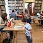 Na zdjęciu widoczni są uczestnicy warsztatów - 4 kobiety i 3 dzieci podczas wykonywania kalendarzy adwentowych z filcu w kształcie Mikołaja. W tle regały z książkami.