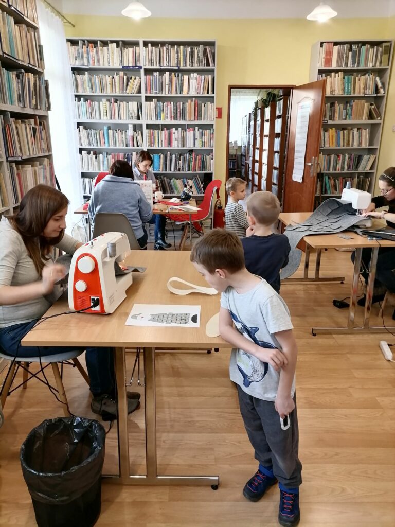 Na zdjęciu widoczni są uczestnicy warsztatów - 4 kobiety i 3 dzieci podczas wykonywania kalendarzy adwentowych z filcu w kształcie Mikołaja. W tle regały z książkami.