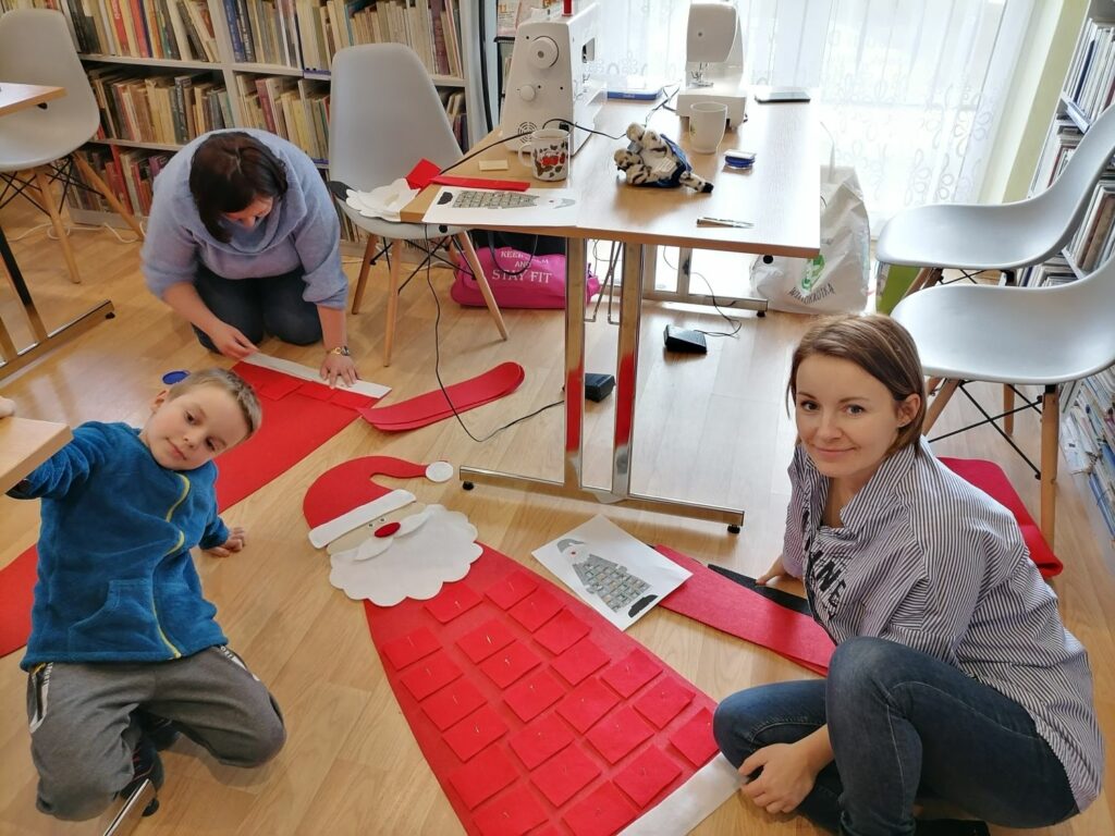 Na zdjęciu widoczni są uczestnicy warsztatów dwie kobiety oraz jedno dziecko - chłopiec, wykonujący kalendarze adwentowe z filcu w kształcie Mikołaja.