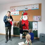 Na zdjęciu stoi bibliotekarz obok niego dziewczynka, która przypina ozdoby do filcowego Mikołaja, który przymocowany jest do tablicy korkowej.