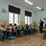 W szkolnej klasie uczniowie siedzą w ławkach i z zainteresowaniem słuchają jak bibliotekarz czyta im książkę pod tytułem „Święta na świecie”.
