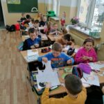 Grupa dzieci siedzi w ławkach i wycina z papieru, a następnie koloruje kredkami ozdobne bombki na choinkę.