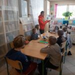 W czytelni przy długim stole siedzi grupa dzieci i z zainteresowaniem słucha czytanej im przez bibliotekarza książki o „Niedoskonałym pingwinie” Kubusiu.