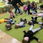 na zdjęciu widać salę przedszkolną, zielona wykładzinę na której leżą dzieci oraz panie opiekunki które im pomagają wykonać polecenia gimnastyczne. Widać również mebelki: krzesła i stoliki.