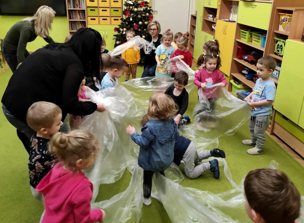 na fotografii widać grupę dzieci. Przedszkolaki bawią się dużą folią malarską. Widać również panie opiekunki oraz choinkę.