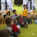 Na fotografii widać dzieci siedzące na zielonej wykładzinie. Jedno z dzieci (chłopiec) stoi przed bibliotekarką i odpowiada na zagadkę.
