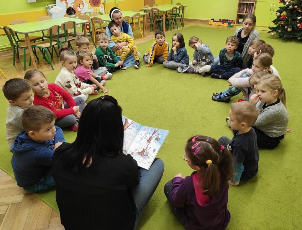 Na fotografii widać dzieci siedzące w kole na zielonej wykładzinie. Dzieci słuchają czytającej bibliotekarki. Na zdjęciu widać wśród dzieci siedzącą opiekunkę, jest to siostra zakonna w habicie.