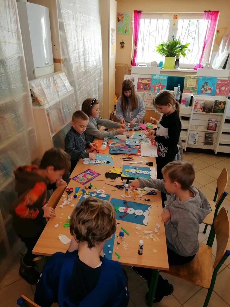 Dzieci pracują przy stole z wielkim zapałem nad swoimi kolorowymi bałwankami, wszędzie pełno jest rozłożonych przyborów plastycznych.