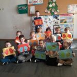 Grupa przedszkolaków ustawiła się do wspólnego zdjęcia. Uśmiechnięte dzieci z dumą prezentują wykonane przez siebie prace plastyczne.