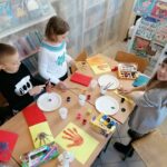 W bibliotecznej sali kilkoro dzieci stoi przy długim i zapełnionym pomocami plastycznymi stole i maluje sobie dłonie farbami.