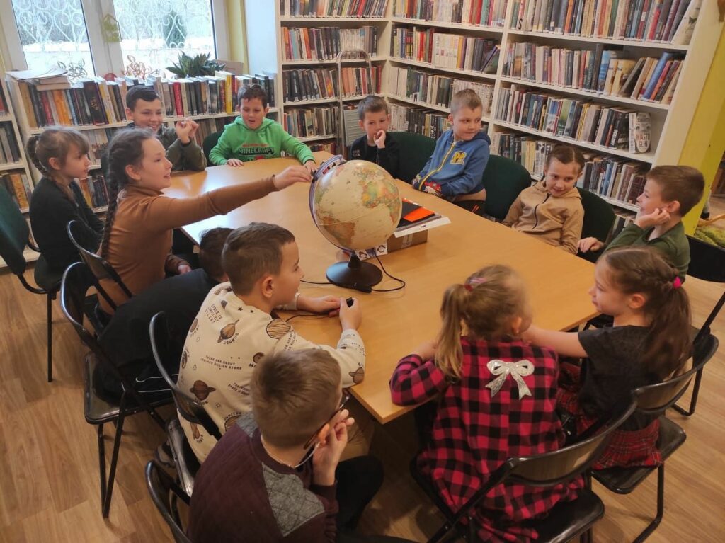 na zdjęciu widać dzieci siedzące przy stole, wykonujące pracę plastyczną na zdjęciu widać salę biblioteczna, regały z książkami, dzieci siedzą wokół stołu, na stole znajduje się globus
