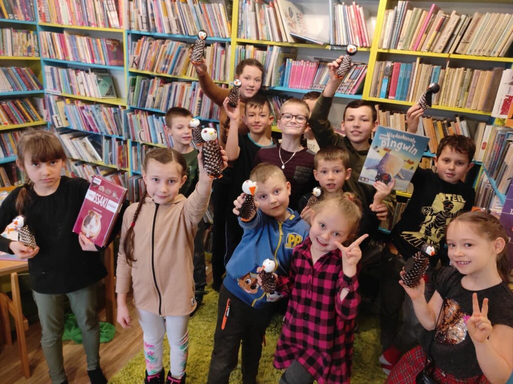 na zdjęciu widać sale biblioteczną, regały z książkami, na środku sali grupa dzieci trzymająca w rękach prace plastyczne