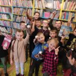 na zdjęciu widać sale biblioteczną, regały z książkami, na środku sali grupa dzieci trzymająca w rękach prace plastyczne
