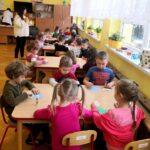 na zdjęciu widać dzieci siedzące w sali przedszkolnej przy stolikach i wykonujące prace plastyczną.
