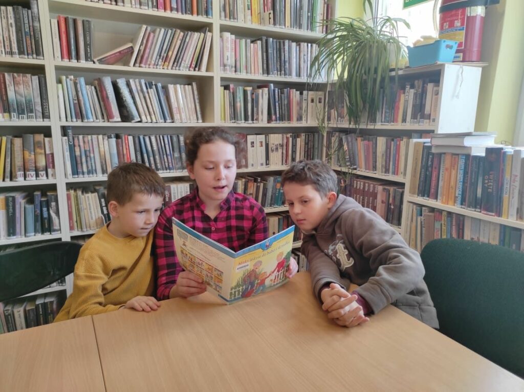 na zdjęciu widać troje dzieci siedzących przy stole, dziewczynka czyta książkę a obok niej z lewej i z prawej strony siedzą chłopcy