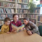 na zdjęciu widać troje dzieci siedzących przy stole, dziewczynka czyta książkę a obok niej z lewej i z prawej strony siedzą chłopcy