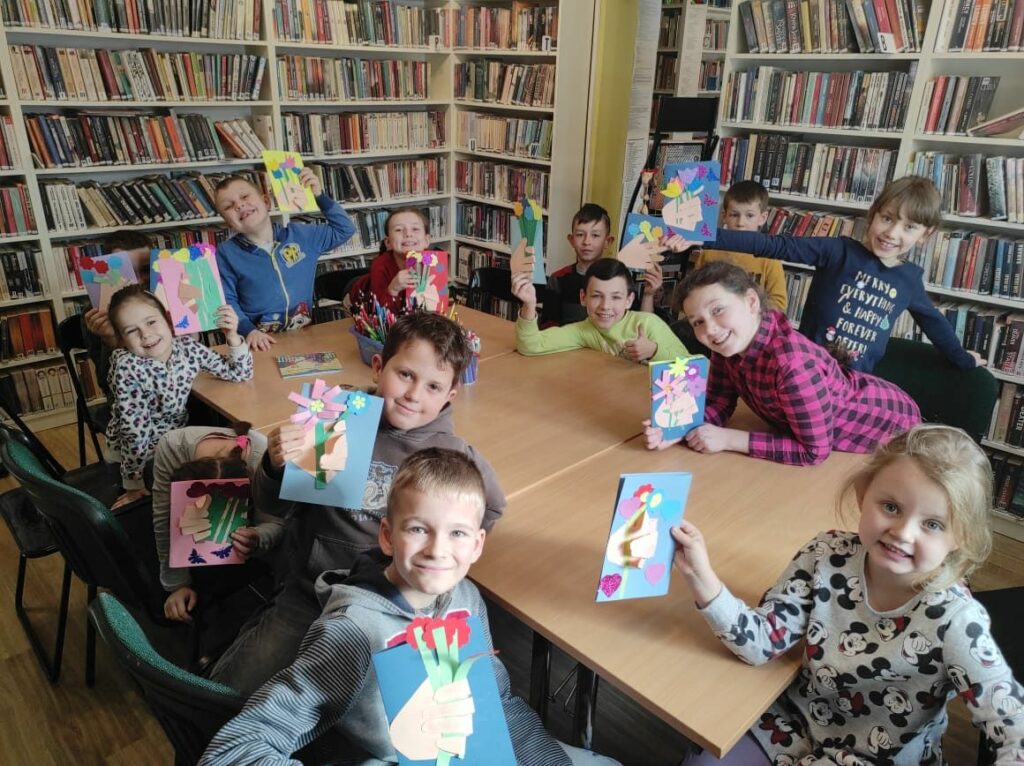 na zdjęci widać salę biblioteczna, regały z książkami, na środku sali stoi stół wokół którego siedzą dzieci, dzieci trzymają w rękach wykonane prace plastyczne - laurki