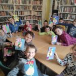 na zdjęci widać salę biblioteczna, regały z książkami, na środku sali stoi stół wokół którego siedzą dzieci, dzieci trzymają w rękach wykonane prace plastyczne - laurki