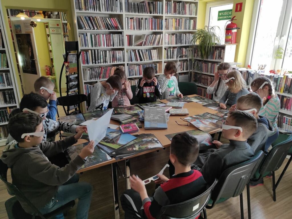 na zdjęciu widać salę biblioteczną, regały z książkami, na środku sali znajduje się stół, wokół stołu siedzą dzieci, mają włożone okulary 3D, na stole leża zdjęcia