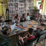 na zdjęciu widać salę biblioteczną, regały z książkami, na środku sali znajduje się stół, wokół stołu siedzą dzieci, mają włożone okulary 3D, na stole leża zdjęcia