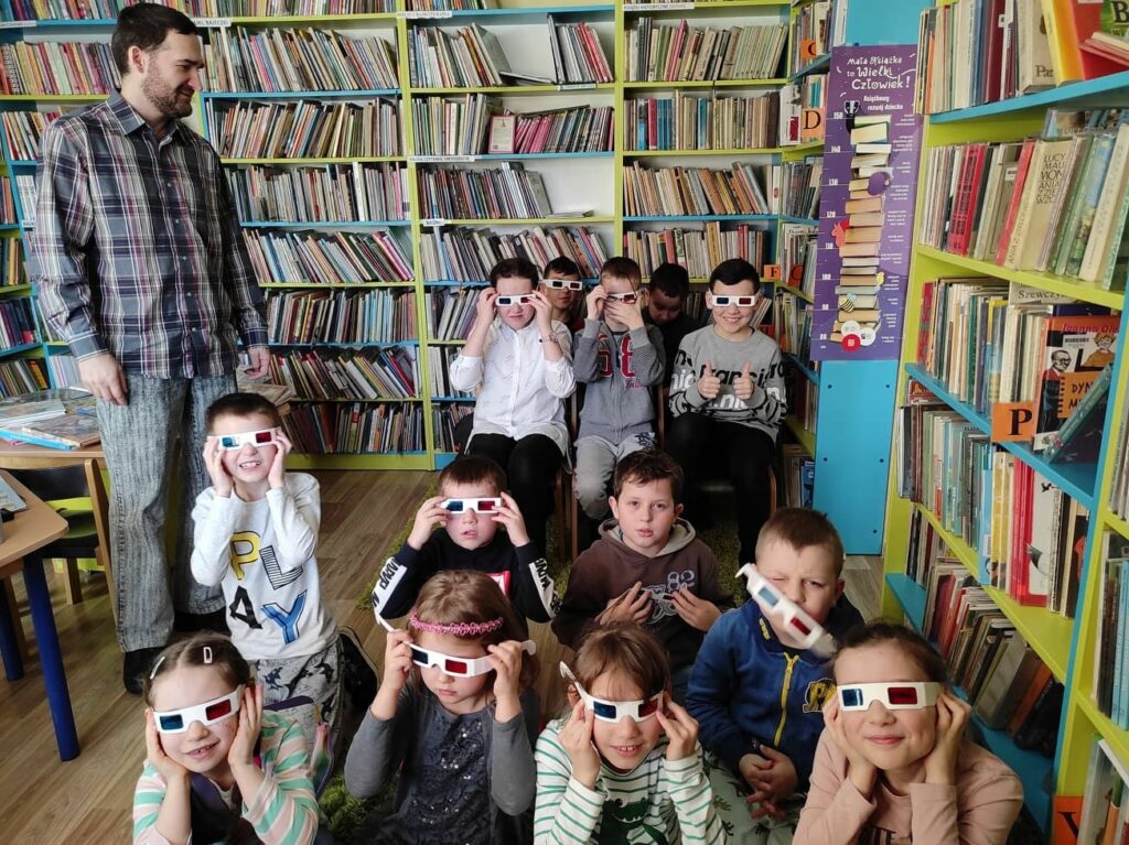 na zdjęciu widać salę biblioteczną, regały z książkami, dzieci siedzą na dywanie albo na krzesłach, dzieci maja włożone okulary 3D, z lewej strony zdjęcia stoi bibliotekarz