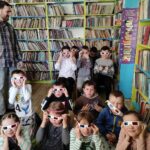 na zdjęciu widać salę biblioteczną, regały z książkami, dzieci siedzą na dywanie albo na krzesłach, dzieci maja włożone okulary 3D, z lewej strony zdjęcia stoi bibliotekarz