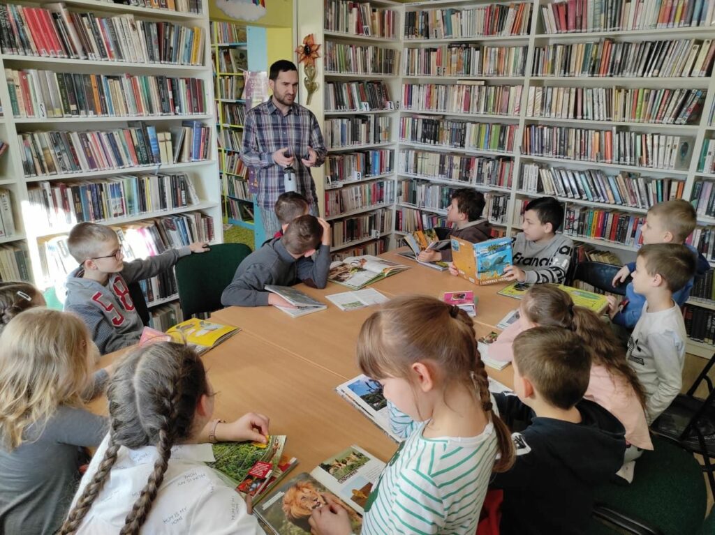 na zdjęciu widać salę biblioteczną, regały z książkami, na środku sali znajduje się stół, wokół stołu siedzą dzieci, każde dziecko ma przed sobą książkę, obok stołu stoi bibliotekarz