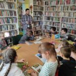 na zdjęciu widać salę biblioteczną, regały z książkami, na środku sali znajduje się stół, wokół stołu siedzą dzieci, każde dziecko ma przed sobą książkę, obok stołu stoi bibliotekarz