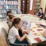 Cztery dziewczynki siedzą przy stole w czytelni biblioteki w Tyliczu. Jedna z dziewczynek czyta książkę.