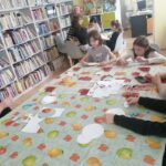 Ptzy dużym stole siedzi piątka dzieci i wycina kółka z papieru technicznego. W dali siedzą cztery dziewczynki przy stoliku i graja w gry. Po lewej stronie regały z książkami.