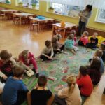 na zdjęciu widać salę przedszkolna, okna, stoliki, krzesełka, parkiet oraz dzieci siedzące w kole, słuchają bibliotekarkę która stoi i czyta im książkę.
