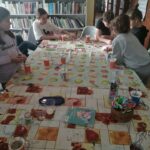 Na zdjęciu w sali bibliotecznej przy dużym stole siedzą dzieci i wykonują prace plastyczne. Na stole położone nożyczki kolorowe, kubeczki.