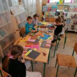W bibliotecznej sali grupa dzieci pracuje przy długim stoliku wycinając z kolorowego papieru swoje zaśnieżone budynki.