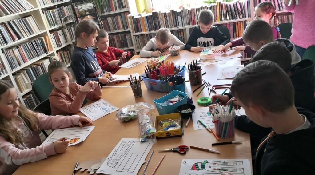 na zdjęciu widać dzieci siedzące przy stole, na którym widać rożnego rodzaju materiały plastyczne np. kredki, nożyczki, pisaki. dzieci kolorują rysunki.