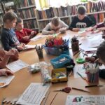 na zdjęciu widać dzieci siedzące przy stole, na którym widać rożnego rodzaju materiały plastyczne np. kredki, nożyczki, pisaki. dzieci kolorują rysunki.