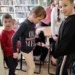 na zdjęciu widać dzieci stojące koło półki, dziewczynka stoi na papierowych rokach papieru, próbując zachować równowagę.