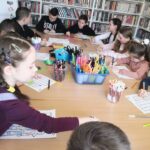 na zdjęciu widać dzieci siedzące przy stole, na którym widać rożnego rodzaju materiały plastyczne np. kredki, nożyczki, pisaki. dzieci kolorują rysunki.