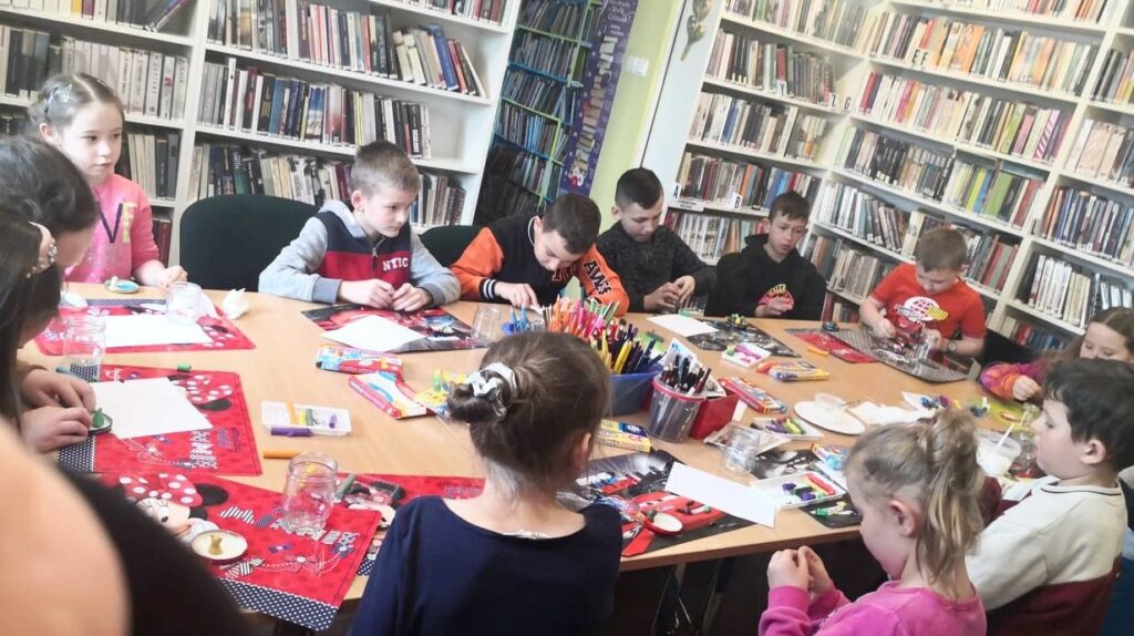 na zdjęciu widać stół w sali biblioteczne wokół którego siedzą dzieci i wykonują prace plastyczną. Na stole kolorowe poskładki, kredki w pudełkach oraz brokat i plastelina