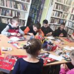 na zdjęciu widać stół w sali biblioteczne wokół którego siedzą dzieci i wykonują prace plastyczną. Na stole kolorowe poskładki, kredki w pudełkach oraz brokat i plastelina