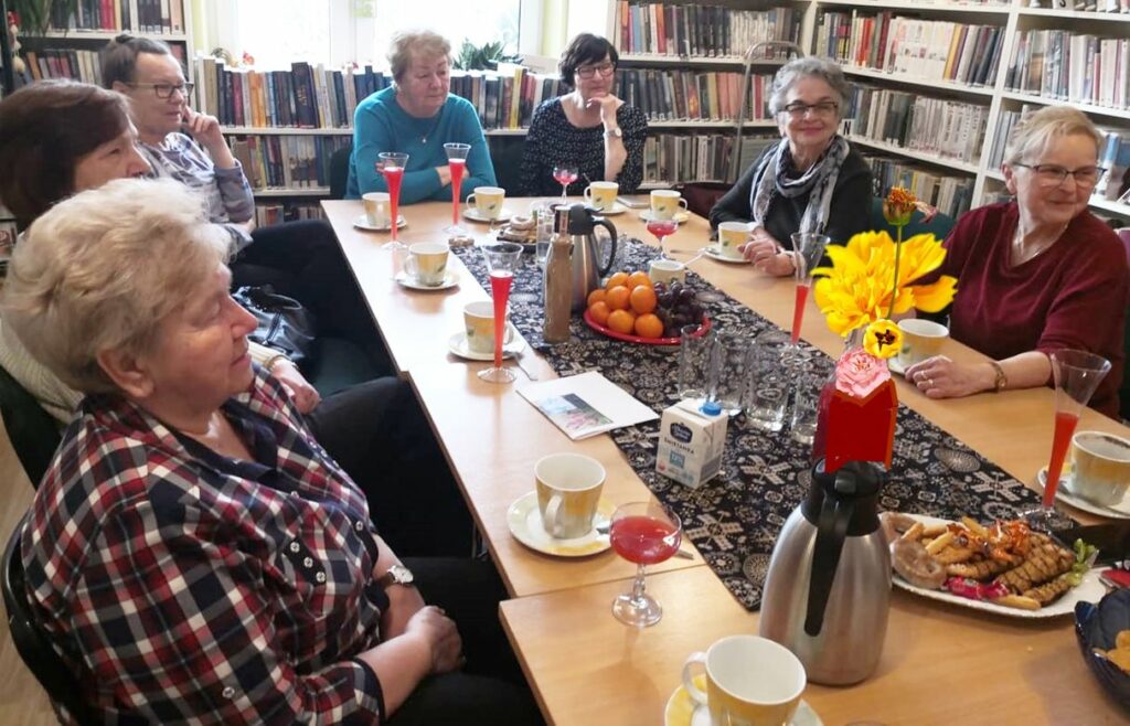 na zdjęciu widać sale biblioteczną, regały z książkami oraz stół przy którym siedzą panie seniorki. Na stole widać kwiaty, filiżanki z kawą oraz szklanki