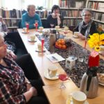 na zdjęciu widać sale biblioteczną, regały z książkami oraz stół przy którym siedzą panie seniorki. Na stole widać kwiaty, filiżanki z kawą oraz szklanki