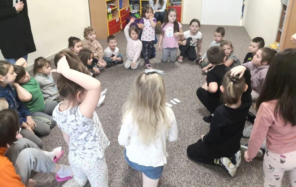 na fotografii widać salę przedszkolną oraz dzieci jedne siedzą inne stoją na beżowej wykładzinie