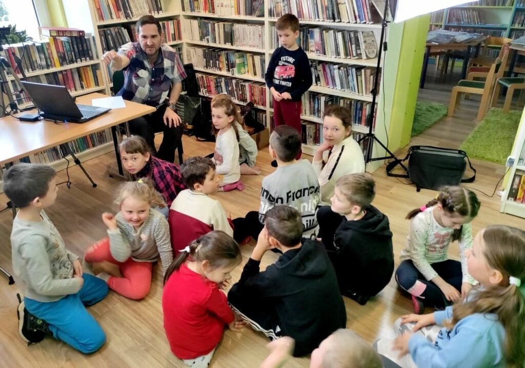 na widać dzieci siedzące na podłodze, salę biblioteczna, stół na stole laptop i siedzący przy nim bibliotekarz objaśniający coś dzieciom