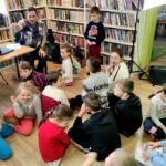 na widać dzieci siedzące na podłodze, salę biblioteczna, stół na stole laptop i siedzący przy nim bibliotekarz objaśniający coś dzieciom
