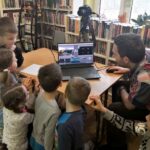 na fotografii widać salę biblioteczną, regały z książkami, stół dzieci wpatrzone w laptop, na którym wyświetlany jest film