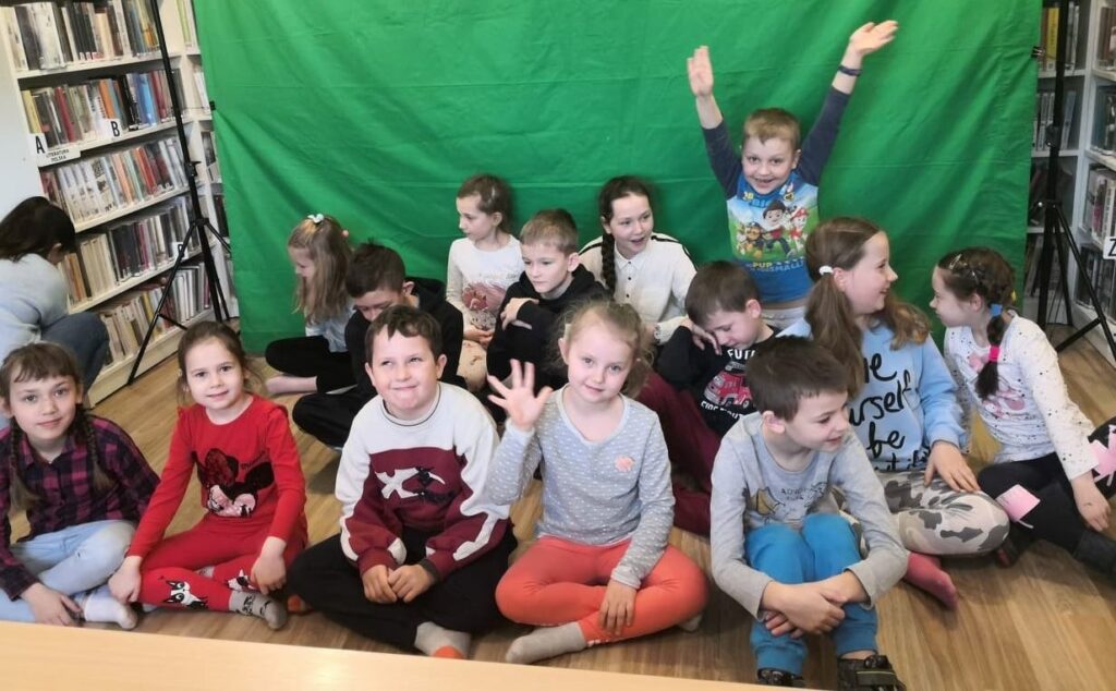 na fotografii widać dzieci siedzące na podłodze na tle zielnego płótna